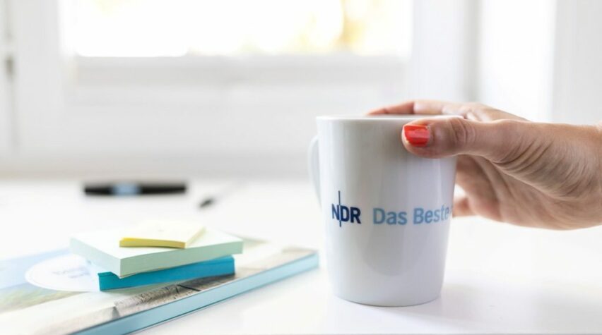 Fotografie einer Kaffeetasse mit NDR Schriftzug und einer Hand.
