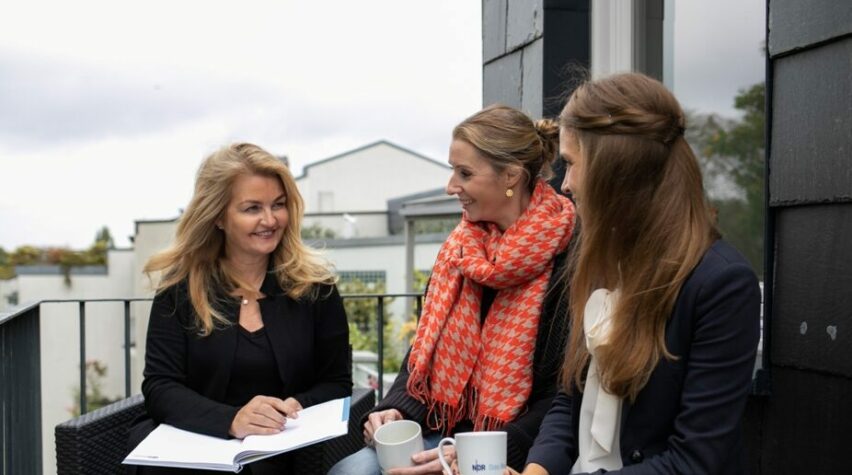 Drei Frauen sitzen auf einem Balkon mit Notizbuch und Kaffeetassen in der Hand.