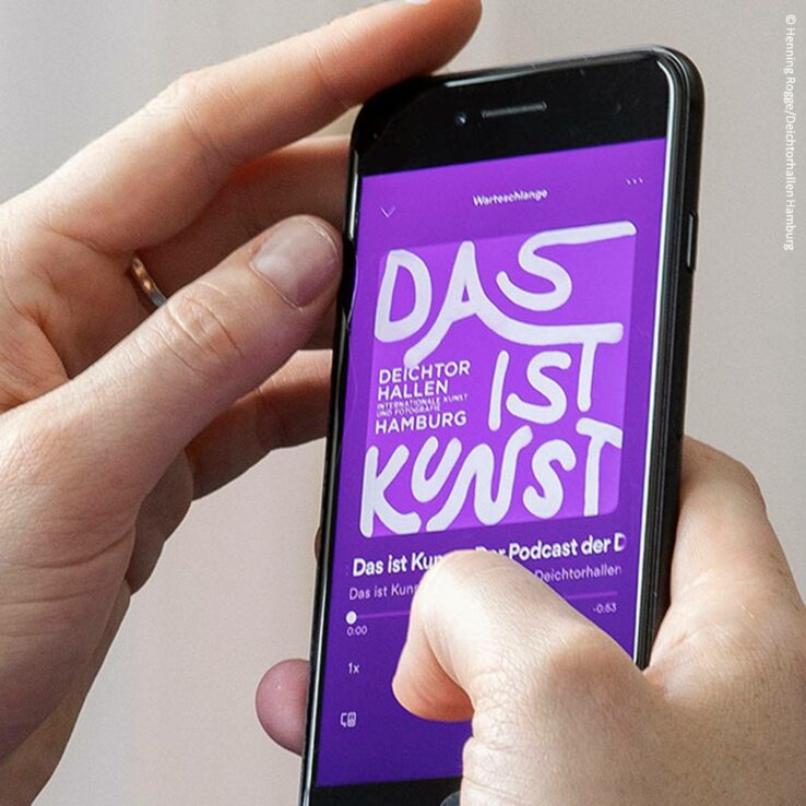 Ein Handy wird in zwei Händen gehalten, darauf spielt eine Audiodatei und der Titel "Das ist Kunst" ist in Weiß auf lila Hintergrund zu sehen.