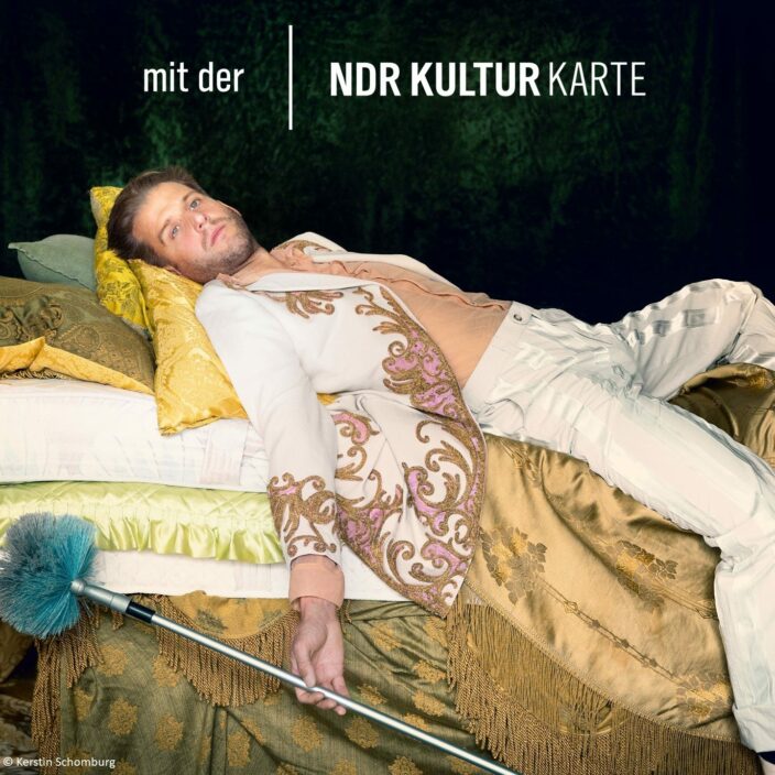 Ein Schauspieler extravangant auf einem Bett, in der Hand ein Staubwedel. Schauspiel Hannover